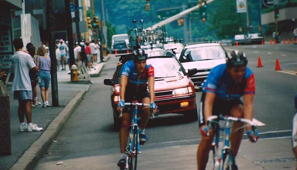 Frankie Andreu racing in a cycling event on a city street with a team car behind him